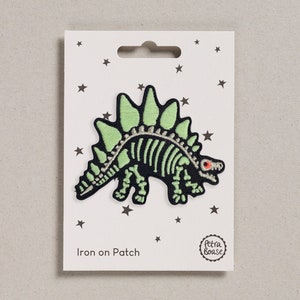 Può includere: Toppa da stirare con uno scheletro di dinosauro verde con occhi rossi. La toppa è su una carta bianca con stelle nere e il testo "Iron on Patch" e "Petra Boase".
