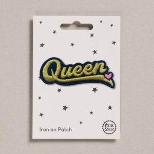 May include: An iron-on patch with the word "Queen" in gold script, outlined in navy blue, and a pink heart. The patch is on a white card with black star designs and the text "Iron on Patch".