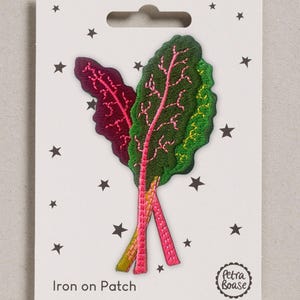 May include: An embroidered iron-on patch featuring two chard leaves with dark green and deep purple foliage, bright pink stems, and yellow accents. The patch is mounted on a white backing card decorated with small black stars and the text Iron on Patch.