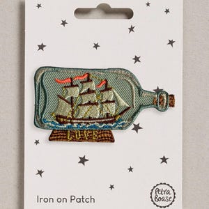 May include: An embroidered iron-on patch featuring a sailing ship inside a glass bottle. The ship has white sails and red flags, resting on a brown wooden base with the word LOVE stitched in gold. The patch is mounted on a white card with stars.
