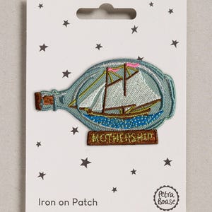May include: An embroidered iron-on patch featuring a ship inside a glass bottle. The ship has white sails, brown masts, and pink flags, resting on blue water. A brown base below the bottle displays the text MOTHERSHIP in yellow embroidery.