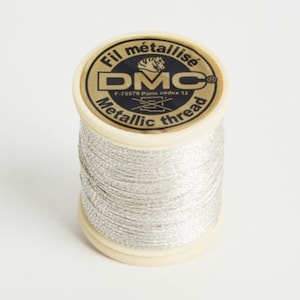Puede incluir: Una bobina de hilo metálico plateado con una etiqueta dorada que dice "Fil metallise DMC Metallic thread F-75579 Paris cedex 12"