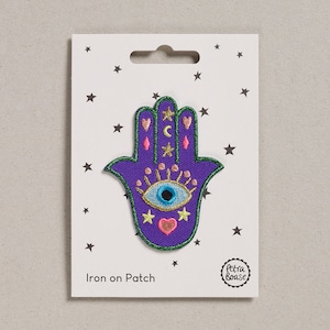May include: A purple Hamsa hand iron-on patch with a green border, featuring an eye, stars, and crescent moons. The patch is on a white card with a star pattern and the text "Iron on Patch".