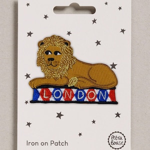 May include: An embroidered iron-on patch featuring a golden lion lying on a rectangular base with a red, white, and blue diamond pattern. The word LONDON is stitched in red letters across the base. The patch is displayed on a white card with stars.