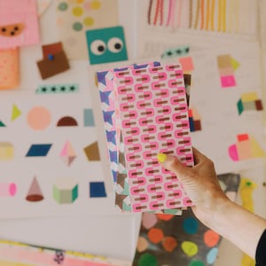 Op de afbeelding: Een stapel notitieboekjes met patronen in roze, bruin en blauw. De notitieboekjes worden vastgehouden tegen een witte achtergrond met kleurrijke geometrische vormen en andere decoratieve elementen.