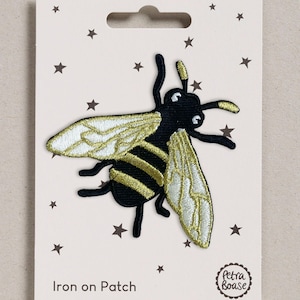 May include: An embroidered iron-on patch featuring a bee with a black body, gold stripes, and shimmering white wings. The patch is attached to a cream-coloured backing card decorated with small brown stars and the text Iron on Patch and Petra Boase.