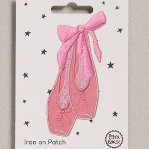 May include: An embroidered iron-on patch featuring a pair of pink ballet slippers with tied ribbons, displayed on a white backing card decorated with small black stars and the text Iron on Patch and the brand name Petra Boase.