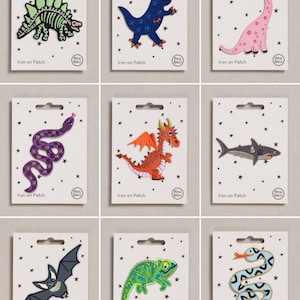 May include: A collection of nine individual iron-on patches displayed on white backing cards with black star patterns. The patches feature embroidered designs of a skeleton dinosaur, blue dinosaur, pink dinosaur, purple snake, orange dragon, shark, bat, green chameleon, and a light blue snake.