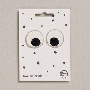 May include: Two round, white fabric patches with black pupils and silver trim, resembling eyes, are displayed on a white card with a star pattern. The card reads "Iron on Patch" and features a logo. These patches are ideal for adding a whimsical touch to clothing or accessories.