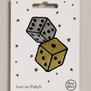 May include: An embroidered iron-on patch featuring two dice, one in metallic silver and one in metallic gold, both with black dots. The patch is displayed on a white backing card decorated with small grey stars and the text Iron on Patch and Petra Boase.