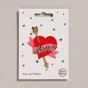 Puede incluir: Parche para planchar con un corazón rojo con una cinta rosa que dice "Love" y una flecha que lo atraviesa. El parche está en una tarjeta blanca con un patrón de estrellas negras.
