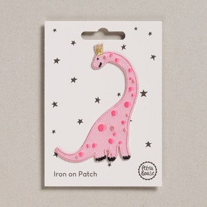 May include: Pink iron-on patch featuring a cartoon dinosaur wearing a gold crown. The dinosaur has pink spots and black feet. The patch is on a white card with black stars and the text "Iron on Patch".