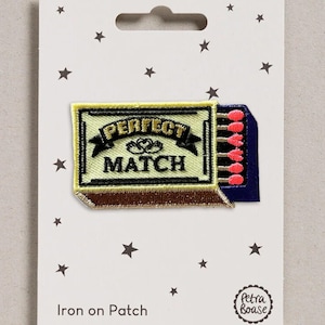 May include: An embroidered iron-on patch shaped like a matchbox with a pale yellow cover, brown base, and dark blue side. The text reads PERFECT MATCH with a small heart icon. Five pink-tipped matches are visible extending from the side of the box.