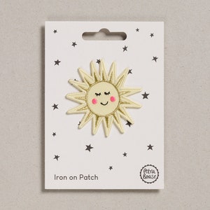 May include: Iron-on patch featuring a yellow sun with a smiling face and pink cheeks. The sun has a gold glitter outline and is on a white card with black stars. The card also says "Iron on Patch" and has a small circle with the text "Petra Boase".