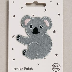 May include: A grey koala iron-on patch with pink inner ears, black eyes, and a black nose. The patch is mounted on a white rectangular card featuring small black stars, the text "Iron on Patch," and a circular "Petra Boase" brand logo.