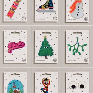 May include: A collection of nine embroidered iron-on patches on white backing cards with black star patterns. Designs include a pink cracker, ice skate, snowman, the word Magic, Christmas tree, mistletoe, snow globe, nutcracker, and googly eyes.