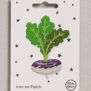 Op de afbeelding: Een geborduurde strijkpatch in de vorm van een knolraap met een paars-witte wortel en felgroene bladeren. De patch is bevestigd op een witte kaart versierd met kleine zwarte sterretjes, de tekst Iron on Patch en het logo van Petra Boase.
