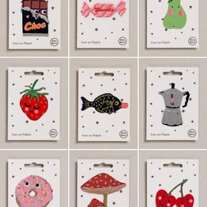 May include: A collection of nine embroidered iron-on patches on white backing cards with black star patterns. Designs include a chocolate bar, candy, pear, strawberry, fish, coffee maker, donut, mushrooms, and cherries, each with embroidered details.