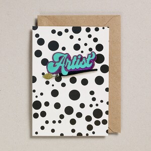 May include: A white card with black polka dots and a turquoise and purple patch that says "Artist" with a gold paintbrush.