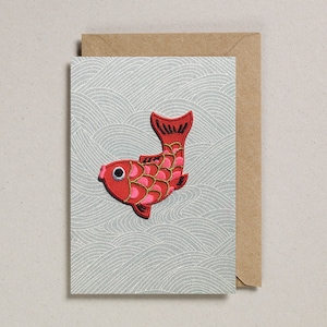 Peut inclure: Un poisson rouge et rose brodé avec des yeux noirs et un contour doré sur une carte bleu clair avec un motif de vagues. La carte est dans une enveloppe brune.