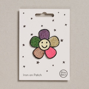 Könnte beinhalten: Bügeleisen-Patch mit einer bunten Blume mit einem lächelnden Gesicht. Die Blume hat Blütenblätter in den Farben Lila, Grün, Gold, Braun und Pink. Der Patch befindet sich auf einer weißen Karte mit Sternen und dem Text "Iron on Patch".