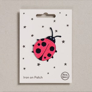 Könnte beinhalten: Bügeleisen-Aufnäher mit einer pinkfarbenen Marienkäfer mit schwarzen Punkten und schwarzen Antennen. Der Aufnäher befindet sich auf einer weißen Karte mit einem schwarzen Sternenmuster und dem Text "Iron on Patch" und "Petra Bouse".