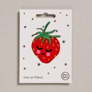May include: A red strawberry iron-on patch with a smiling face, closed eyes, and pink cheeks. The patch has green leaves and is on a white card with black stars. The text "Iron on Patch" is printed on the card.