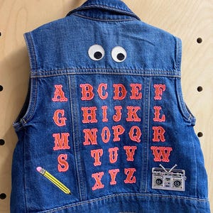 May include: A blue denim vest with a playful design. The back features large, red embroidered letters of the alphabet, googly eyes, a pencil, and a boombox patch. The vest is hanging on a wooden hanger.