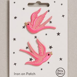 May include: Two embroidered iron-on patches shaped like swallows in bright pink with gold-coloured beaks, displayed on a white backing card decorated with black stars and the text Iron on Patch and the brand name Petra Boase.
