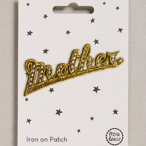 Op de afbeelding: Een opstrijkbare patch met het woord mother in gouden metallic draad met witte accenten. De patch is bevestigd op een witte rechthoekige kaart versierd met kleine zwarte sterren, de tekst Iron on Patch en een rond Petra Boase logo.