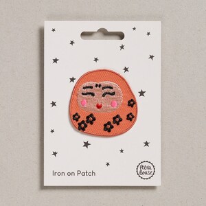 May include: Iron-on patch featuring a smiling orange daruma doll with black eyes and a pink mouth. The doll is surrounded by black flowers. The patch is on a white card with the text "Iron on Patch" and a logo that says "Petra Boase".