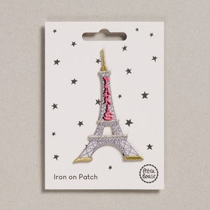 May include: Iron-on patch featuring a silver Eiffel Tower with a pink "PARIS" inscription and gold accents. The patch is on a white card with black stars and the text "Iron on Patch" and "Petra Boase".