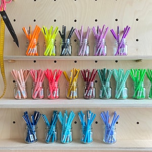 Könnte beinhalten: Eine Sammlung farbiger Stifte in kleinen Gläsern, angeordnet auf Holzregalen. Die Stifte sind in verschiedenen Farben erhältlich, darunter Orange, Gelb, Schwarz, Rosa, Lila, Rot, Grün, Blau und Hellblau. Eine Schere und ein Maßband sind links zu sehen.