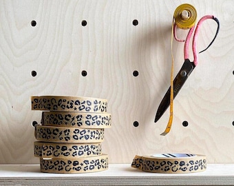 May include: A stack of five rolls of brown washi tape with a black leopard print and white stars. The tape is on a wooden shelf with a yellow measuring tape and pink scissors.