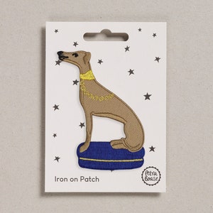 Op de afbeelding: Strijk-op patch met een bruine hond die een gouden halsband draagt en op een blauw kussen zit. De patch heeft een witte achtergrond met sterren en de tekst "Iron on Patch" en "Petra Boase" in zwart.