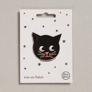 May include: Iron-on patch featuring a black cat with pink nose and yellow whiskers. The patch is on a white card with black stars and the text "Iron on Patch" and a logo that says "Petra Boase".