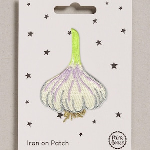 Op de afbeelding: Een geborduurde strijkpatch in de vorm van een knoflookbol met een lichtgroene stengel, lichtpaarse en crèmekleurige teentjes en goudkleurige wortels. De patch is bevestigd op een witte kaart met zwarte sterren en de tekst Petra Boase Iron on Patch.