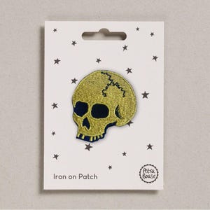 Peut inclure: Écusson thermocollant en forme de crâne brodé en or avec des détails bleu marine. L'écusson est sur une carte blanche avec un motif d'étoiles et le texte "Iron on Patch".