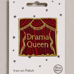 May include: A square iron-on patch featuring an embroidered design of deep red stage curtains with gold trim and tassels. The text "Drama Queen" is stitched in gold thread in the centre. The patch is mounted on a white card with black stars.