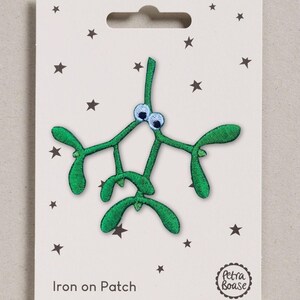 May include: An embroidered iron-on patch shaped like a green mistletoe sprig with two large googly eyes. The patch is attached to a white rectangular display card featuring small black stars, the text Iron on Patch, and a Petra Boase logo.