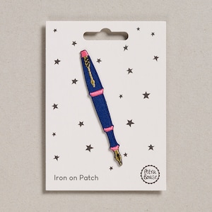 May include: A navy blue iron-on patch in the shape of a fountain pen, featuring pink and gold detailing. The patch is displayed on a white card adorned with star motifs and the text "Iron on Patch". The Petra Boase logo is visible.