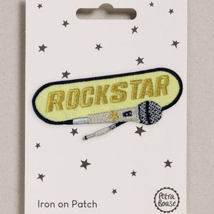 May include: An iron-on patch featuring a pale yellow oval background with a black border and the word ROCKSTAR embroidered in gold thread. A silver and black microphone with a small gold star detail is positioned below the text.