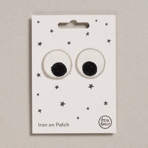 May include: Two white iron-on patches with black eyes and a silver outline. The patches are on a white card with a small star pattern and the text "Iron on Patch" and a logo that says "Petra Boase".