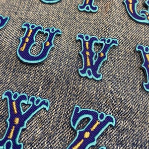 May include: Close-up of embroidered fabric letters in a decorative font. The letters are dark blue with a light blue outline and gold accents. The letters are arranged on a denim background.