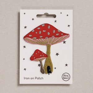 May include: Iron-on patch featuring two embroidered mushrooms. The larger mushroom is red with white polka dots and a pink underside. The smaller mushroom is red with white polka dots. Both mushrooms have gold stems and a gold door on the larger mushroom. The patch is on a white card with the text "Iron on Patch" and a small logo that says "Petra Boase".
