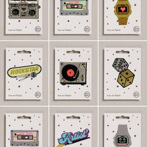 May include: A collection of nine embroidered iron-on patches displayed on individual white cards with star patterns. The designs include a boombox, cassette tapes, a digital watch, a rockstar sign, a record player, dice, the word Artist, and a watch.