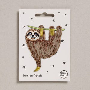 May include: An iron-on patch depicting a brown sloth hanging from a green branch. The sloth has a smiling face and yellow claws. The patch is on a white card with black star accents and the text "Iron on Patch".