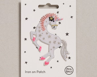 May include: Iron-on patch featuring a white unicorn with pink hooves and a pink mane. The unicorn has a gold horn and a black star on its forehead. The patch is surrounded by white stars on a white background. The text "Iron on Patch" is printed below the unicorn. The text "Petra Boase" is printed in a circle below the text "Iron on Patch".