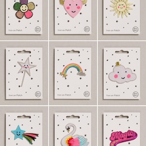 May include: A collection of nine embroidered iron-on patches on white backing cards with black star patterns. Designs include a multi-coloured flower, a winged heart, a sun, a star wand, a rainbow, a cloud, a shooting star, a swan, and the word Magic.