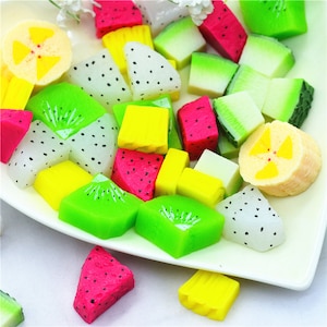 Puede incluir: Una colección de rodajas de frutas artificiales de colores, dispuestas sobre un plato blanco. El surtido incluye kiwi verde, sandía roja, plátano amarillo y fruta del dragón blanca, todos con detalles realistas. Los colores vibrantes crean una exhibición lúdica.